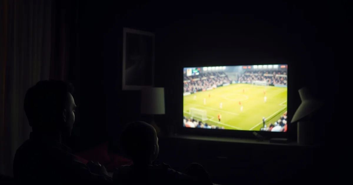 Salon sombre et cinématographique à Dakar : une famille regarde un match de football en direct sur grand écran, la lumière chaude du téléviseur contrastant avec l'obscurité de la pièce, un téléphone allumé à proximité.
